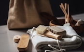 A collection of eco-friendly personal hygiene items is arranged on a soft, white fabric. Included are a wooden brush with natural bristles, a loofah, and other bamboo or wooden accessories, suggesting a focus on sustainability. The scene is softly lit, and a burlap bag is subtly visible in the blurred background.