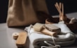 A collection of eco-friendly personal hygiene items is arranged on a soft, white fabric. Included are a wooden brush with natural bristles, a loofah, and other bamboo or wooden accessories, suggesting a focus on sustainability. The scene is softly lit, and a burlap bag is subtly visible in the blurred background.