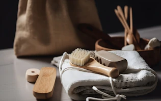 A collection of eco-friendly personal hygiene items is arranged on a soft, white fabric. Included are a wooden brush with natural bristles, a loofah, and other bamboo or wooden accessories, suggesting a focus on sustainability. The scene is softly lit, and a burlap bag is subtly visible in the blurred background.