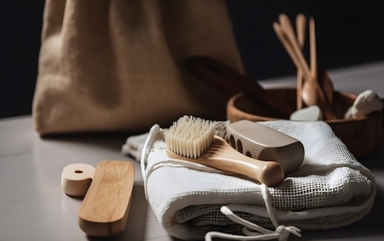 A collection of eco-friendly personal hygiene items is arranged on a soft, white fabric. Included are a wooden brush with natural bristles, a loofah, and other bamboo or wooden accessories, suggesting a focus on sustainability. The scene is softly lit, and a burlap bag is subtly visible in the blurred background.