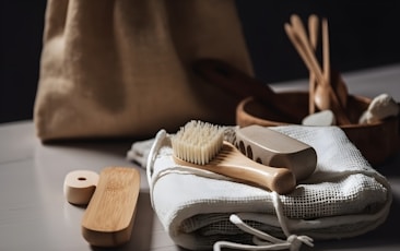 A collection of eco-friendly personal hygiene items is arranged on a soft, white fabric. Included are a wooden brush with natural bristles, a loofah, and other bamboo or wooden accessories, suggesting a focus on sustainability. The scene is softly lit, and a burlap bag is subtly visible in the blurred background.