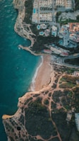 An aerial view of a coastal area with a small sandy beach surrounded by rugged cliffs. Residential buildings with pools are situated on top of the cliffs, with roads and greenery around them. The turquoise sea meets the shore with gentle waves.