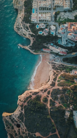 An aerial view of a coastal area with a small sandy beach surrounded by rugged cliffs. Residential buildings with pools are situated on top of the cliffs, with roads and greenery around them. The turquoise sea meets the shore with gentle waves.