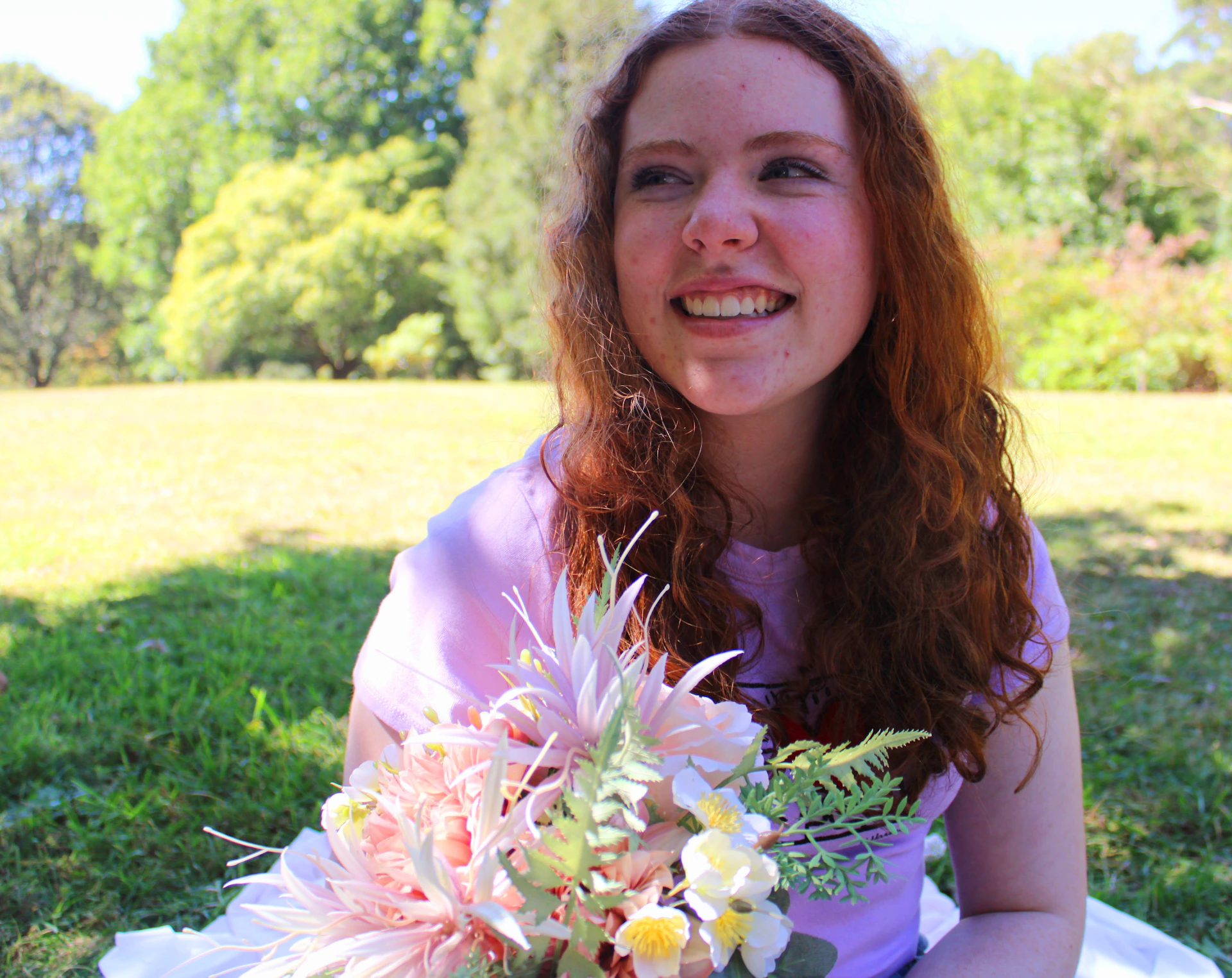A warm candid photo of Agnieszka smiling gently during a sunny afternoon in a park, surrounded by blooming flowers.