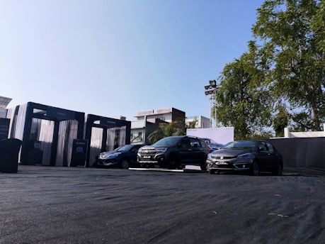 Three cars are parked on a black tarp-covered outdoor space. The area is framed by modern architectural elements and a lush tree on the right. A clear blue sky enhances the bright atmosphere, while a billboard with a car advertisement is visible in the background.