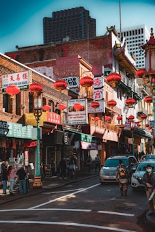 A bustling urban street scene featuring a busy China Town area, with traditional red lanterns hanging between buildings adorned with ornate architectural details and vibrant signage. People walk the street, and cars are parked along the side.