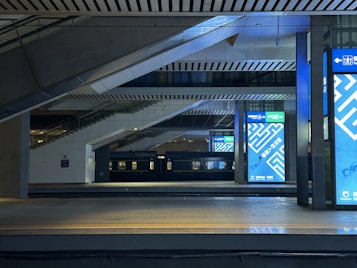 A modern train station platform with large digital screens displaying advertisements featuring geometric patterns in blue. The architectural design includes angular and linear elements with a combination of several levels and overhead beams. A stationary black train is visible on the adjacent track, and the area is well-lit, highlighting the metallic and concrete surfaces.
