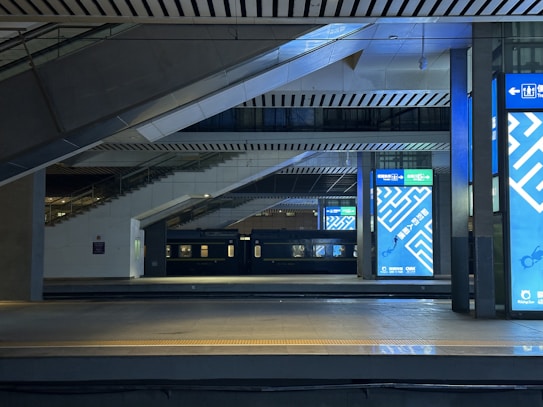 A modern train station platform with large digital screens displaying advertisements featuring geometric patterns in blue. The architectural design includes angular and linear elements with a combination of several levels and overhead beams. A stationary black train is visible on the adjacent track, and the area is well-lit, highlighting the metallic and concrete surfaces.