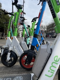 A row of parked electric scooters with varying colors like green, white, and blue stand on a pavement in an urban setting. Buildings and bare trees can be seen in the background, indicating a possible winter or early spring day.