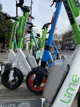 A row of parked electric scooters with varying colors like green, white, and blue stand on a pavement in an urban setting. Buildings and bare trees can be seen in the background, indicating a possible winter or early spring day.