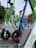 A row of parked electric scooters with varying colors like green, white, and blue stand on a pavement in an urban setting. Buildings and bare trees can be seen in the background, indicating a possible winter or early spring day.