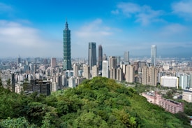 A bustling urban skyline characterized by a mix of modern skyscrapers and mid-rise buildings. The landscape features lush green hills in the foreground with a prominent tall building that stands out, tapering towards the top. The sky is clear with a few clouds.