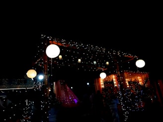 A beautifully decorated outdoor setting with strings of twinkling lights and hanging lanterns creating a warm, festive ambiance. The scene is dimly lit, enhancing the glow of the lights and lanterns, with hints of red and orange from the draped materials.