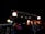 A beautifully decorated outdoor setting with strings of twinkling lights and hanging lanterns creating a warm, festive ambiance. The scene is dimly lit, enhancing the glow of the lights and lanterns, with hints of red and orange from the draped materials.