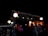 A beautifully decorated outdoor setting with strings of twinkling lights and hanging lanterns creating a warm, festive ambiance. The scene is dimly lit, enhancing the glow of the lights and lanterns, with hints of red and orange from the draped materials.