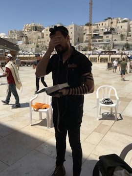 A person with their hand on their forehead appears to be engaged in prayer or a moment of reflection, holding a book. They are wearing a tefillin set, including straps around the arm and a headpiece. The setting is an outdoor plaza with stone buildings in the background. Other people can be seen walking around, some wearing prayer shawls.