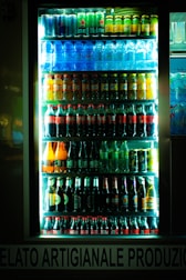 A brightly lit refrigerator display filled with various bottles and cans of beverages, including water, soda, beer, and energy drinks. The drinks are organized in neat rows on multiple shelves inside a glass door cooler.