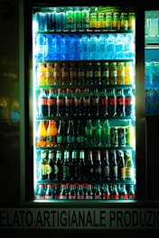 A brightly lit refrigerator display filled with various bottles and cans of beverages, including water, soda, beer, and energy drinks. The drinks are organized in neat rows on multiple shelves inside a glass door cooler.