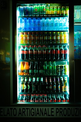 A brightly lit refrigerator display filled with various bottles and cans of beverages, including water, soda, beer, and energy drinks. The drinks are organized in neat rows on multiple shelves inside a glass door cooler.