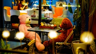 Two large teddy bears are seated at a table in a cozy, dimly lit cafe. The pink and brown bears are positioned as if having a conversation, surrounded by decorative teapots and potted plants. In the background, a woman is visible, engaged in conversation, with colorful chairs and subdued lighting adding to the eclectic and warm atmosphere.