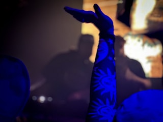 A person raises their arm in a dark, atmospheric setting. The arm is decorated with a sleeve featuring sun and star patterns, illuminated in blue light against a blurred background, possibly suggesting a concert or club environment.
