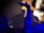 A person raises their arm in a dark, atmospheric setting. The arm is decorated with a sleeve featuring sun and star patterns, illuminated in blue light against a blurred background, possibly suggesting a concert or club environment.