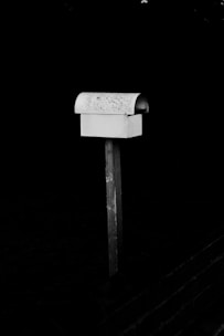 A solitary, weathered mailbox is mounted on a wooden post against a dark background. The mailbox is slightly rusted, with a curved top and rectangular base.