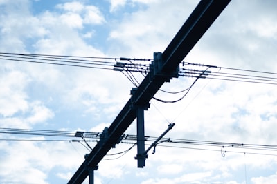A series of electrical power lines and cables are suspended in the air against a backdrop of a partly cloudy blue sky. The cables are supported by a dark metal structure with insulators and connectors visible.