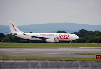 An airplane is taxiing on a runway with the airline branding 'Jet2' prominently displayed on the aircraft's side. The background features a landscape of green trees and distant hills under an overcast sky.