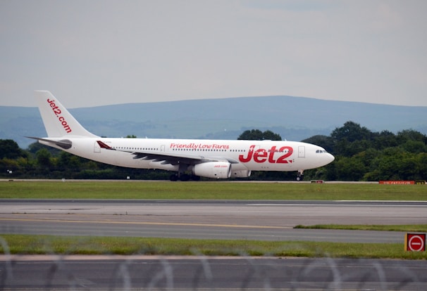 An airplane is taxiing on a runway with the airline branding 'Jet2' prominently displayed on the aircraft's side. The background features a landscape of green trees and distant hills under an overcast sky.