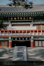 A traditional Korean temple entrance featuring intricate architectural details such as ornate carvings and colorful patterns. Stone steps lead up to wooden doors, and there is a signboard with Korean characters displayed prominently.