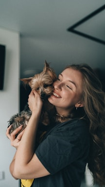 A person with long hair is joyfully holding a small dog close to their face. The person is smiling with eyes closed, expressing affection. The dog appears content and relaxed. The setting seems to be indoors with soft lighting.