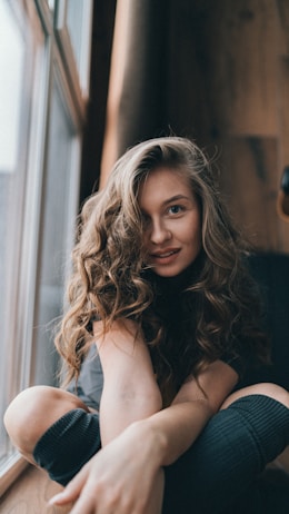 A person with long, curly hair sits comfortably by a window. Sunlight creates a warm ambiance, highlighting the wood-paneled walls in the background. The person's relaxed pose and faint smile suggest a sense of calm and contentment.