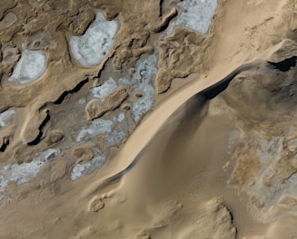 An aerial view of a rugged, desert-like terrain with a mix of sandy and rocky surfaces. Patches of irregular shapes with a lighter color suggest areas that might be dried up or covered with a thin layer of mineral deposits. The image captures a blend of textures, including smooth sandy surfaces and rough, rocky formations.