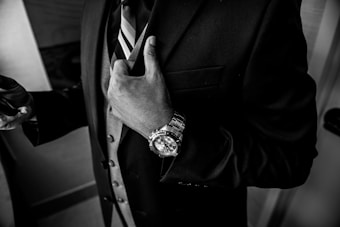 A person dressed in a formal suit with a striped tie is adjusting the jacket. The individual is wearing a luxury watch with a metallic strap on their wrist, and only the torso and hands are visible in the monochrome image.