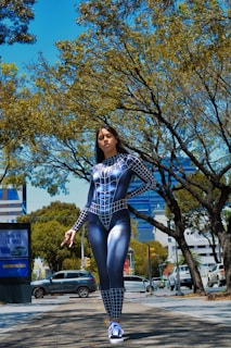 A person dressed in a black and white spider-themed superhero costume stands confidently on a city street. Surrounding the individual are tall trees with green leaves under a bright blue sky. Several vehicles are parked along the road, and there are buildings visible in the background.