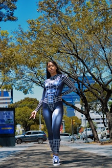 A person dressed in a black and white spider-themed superhero costume stands confidently on a city street. Surrounding the individual are tall trees with green leaves under a bright blue sky. Several vehicles are parked along the road, and there are buildings visible in the background.