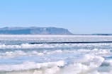 A vast expanse of frozen ice stretches across the foreground, with patches of snow covering the surface. In the distance, low mountains with a dusting of snow and bare ridges rise under a clear blue sky, creating a serene and remote arctic landscape.