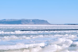 A vast expanse of frozen ice stretches across the foreground, with patches of snow covering the surface. In the distance, low mountains with a dusting of snow and bare ridges rise under a clear blue sky, creating a serene and remote arctic landscape.
