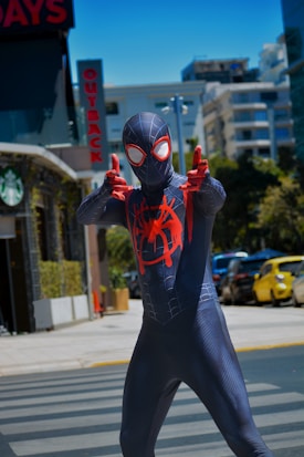 A person wearing a black and red superhero costume poses confidently on a street. The suit features a web-like pattern and a distinctive spider emblem on the chest. The scene includes urban elements such as a 'Starbucks' sign and other city buildings in the background. The lighting suggests it's a sunny day.
