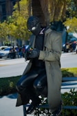 A person wearing a black full-face mask and goggles sits on a railing while solving a Rubik's cube. They are dressed in a long, green trench coat, black leather pants, and gloves. Surrounding them is a cityscape with trees and people walking in the background.
