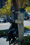 A person wearing a black full-face mask and goggles sits on a railing while solving a Rubik's cube. They are dressed in a long, green trench coat, black leather pants, and gloves. Surrounding them is a cityscape with trees and people walking in the background.