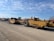 Several large yellow construction vehicles are parked on a dirt lot under a blue sky with scattered clouds. The vehicles are industrial equipment used in earthmoving, with large tires and metal frames.