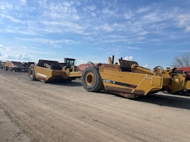 Several large yellow construction vehicles are parked on a dirt lot under a blue sky with scattered clouds. The vehicles are industrial equipment used in earthmoving, with large tires and metal frames.