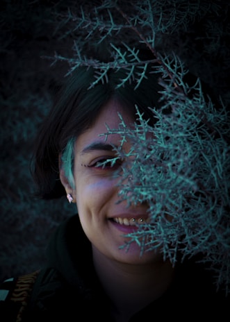 A young adult wearing a pair of green seed studio earrings, their smile soft and genuine against a leafy outdoor backdrop.