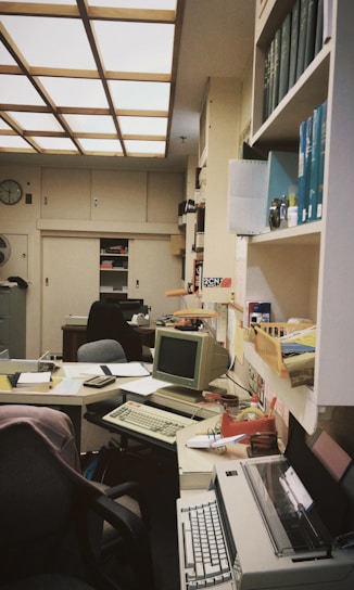 An office space cluttered with various office supplies and equipment. There are several chairs, desks filled with papers, books, and vintage electronics including a CRT monitor, keyboard, and an old typewriter. Shelves filled with binders, books, and file organizers are visible. Lighting is provided by a grid of ceiling panels.