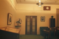 A dimly lit room with vintage decor. A large ceiling light fixture illuminates the space. There's a wooden table or desk visible on the left and a few chairs along the right side. A wall clock hangs above a set of double doors, and a framed photograph is mounted to the right of the doors. A wall-mounted fan is on the left side of the doors, next to a picture. The room has a somewhat nostalgic feel.