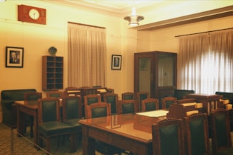 A room with a meeting or conference setup featuring several wooden tables and green padded chairs. The walls are adorned with two portrait photographs, and there is a large wooden clock above the door. The room is softly lit, with curtains covering the windows and a ceiling light fixture providing ambient light.