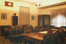 A room with a meeting or conference setup featuring several wooden tables and green padded chairs. The walls are adorned with two portrait photographs, and there is a large wooden clock above the door. The room is softly lit, with curtains covering the windows and a ceiling light fixture providing ambient light.