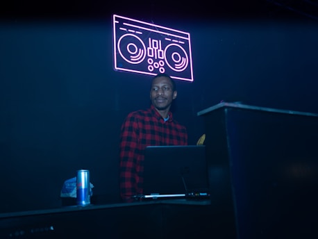 A person wearing a red and black plaid shirt stands behind a DJ booth. A neon sign of a turntable glows above in pink, creating a vibrant atmosphere. A laptop is placed on the booth alongside a can of energy drink.